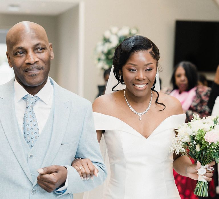 Bride walks down the aisle arm in arm with her father who wears pale blue suit and white shirt