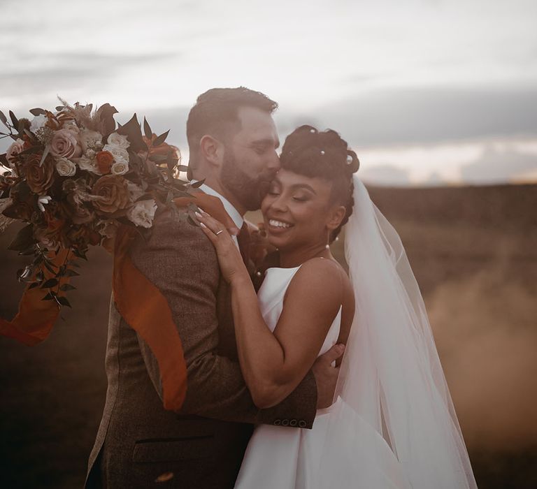 Groom kisses his bride outdoors as she holds dried floral bouquet in Autumnal colours tied with orange sheer ribbon