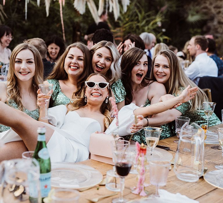 Bride in puff sleeve wedding dress and white sunglasses lounges across the bridesmaids in sequin green dresses