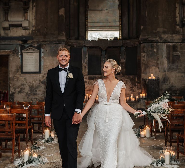 Bride wears low-neck capped sleeve embellished wedding dress whilst holding pampas grass bouquet 