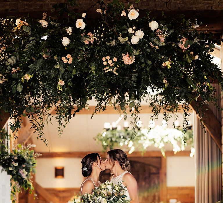 Brides in lace wedding dresses share a kiss holding their bouquets under pink and white flower decor 