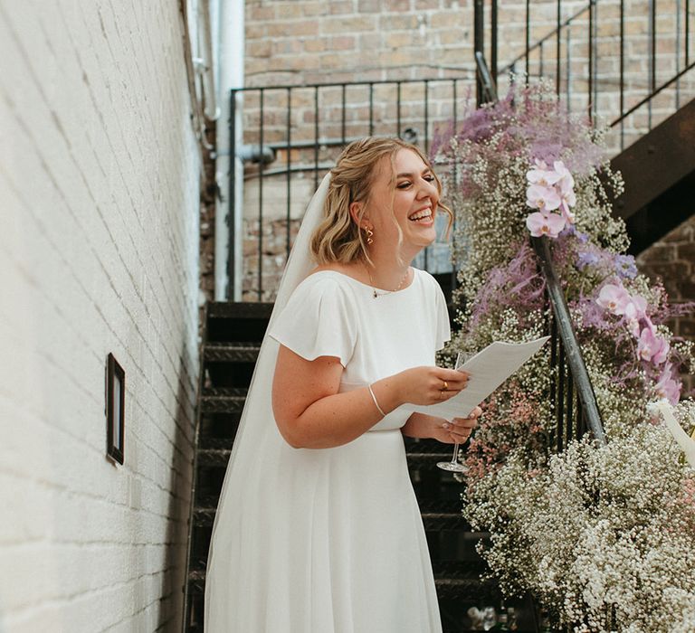 Bride in a short sleeve wedding dress smiling reading out her bridal speech for her wedding at Loft Studios 