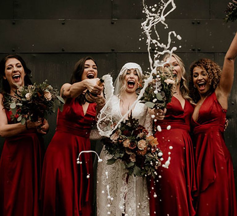 Bride in a lace cap veil and wedding dress popping champagne with the bridesmaids in crimson red magenta dresses 