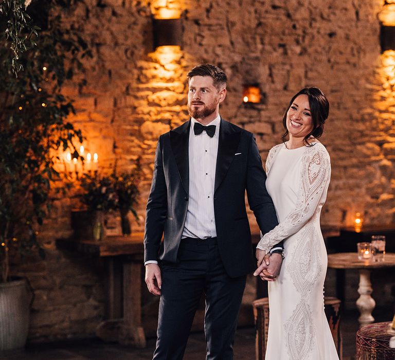 Bride in a long sleeve fitted wedding dress and groom in custom suit stand holding hands together 