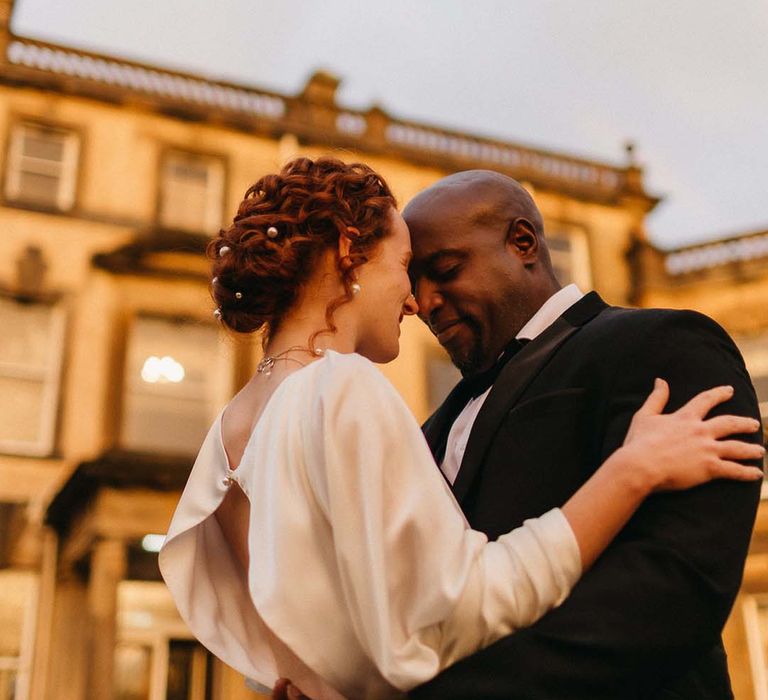 Bride with open back detail wedding dress and stylish up-do with pearls and groom in black tux embracing at Pepper Arden Hall