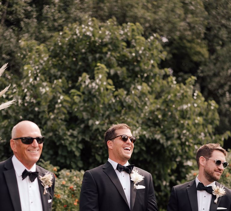 Groom and groomsmen stand together in black tie with sunglasses waiting at the aisle for the bride 