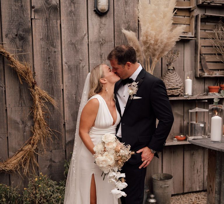 Bride in a satin white wedding dress with a cathedral length veil that has floral embroidery kissing the groom in a black tuxedo for boho luxe wedding 