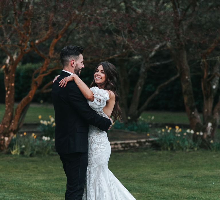 Boho lace strapless wedding dress with detachable sleeves sharing an intimate embrace with the groom in black tie 