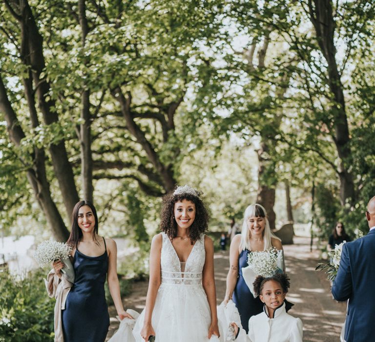 Bride walks alongside her bridesmaids in navy midi bridesmaid dresses and flower girl in white dress with tutu styled skirt