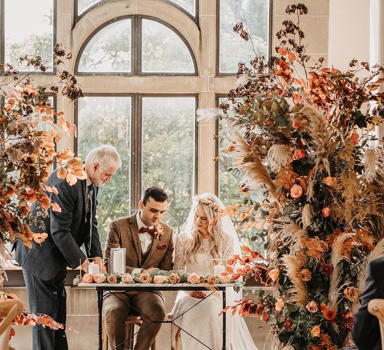 Groom in brown suit signing the register with the bride in a lace dress and flower crown with autumnal flower columns with pampas grass