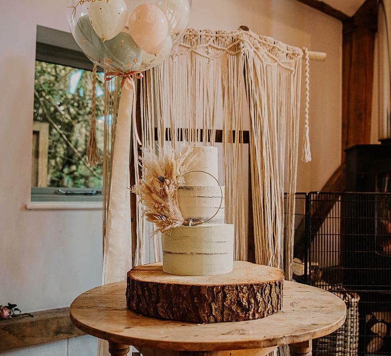 Three tier semi-naked wedding cake on a rustic wooden tree trunk cake stand with a hula hoop cake decorating with pampas grass next to balloon decoration