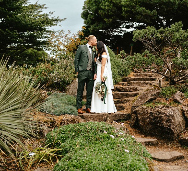 Groom in green three-piece suit and black bow-tie kisses his bride in off-the-shoulder wedding dress during outdoor couples portraits 