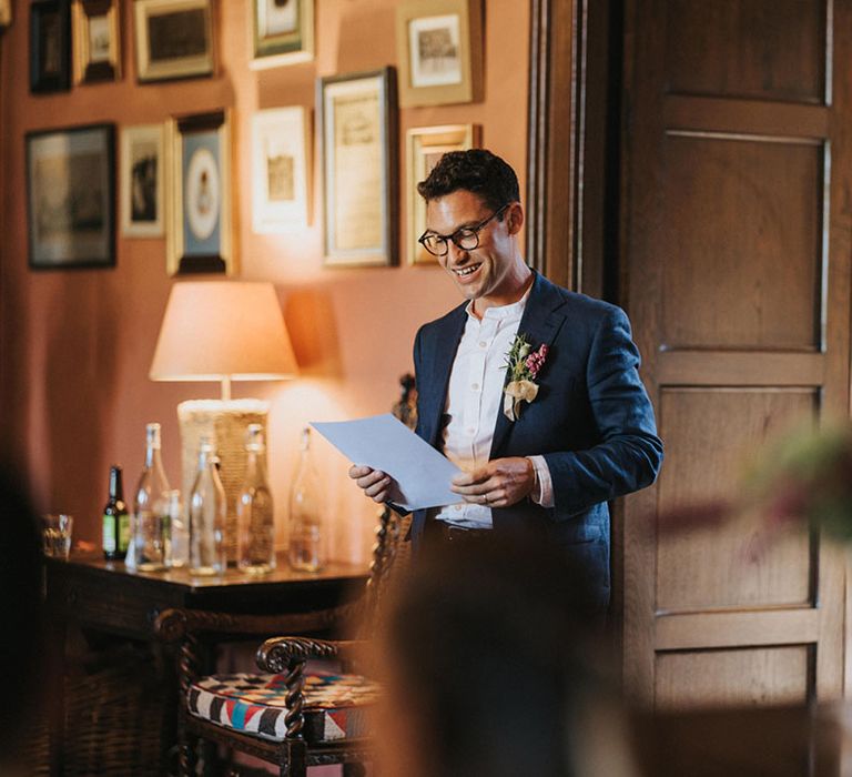 Groom standing in navy wedding suit with plain white shirt as he reads out his wedding speech 