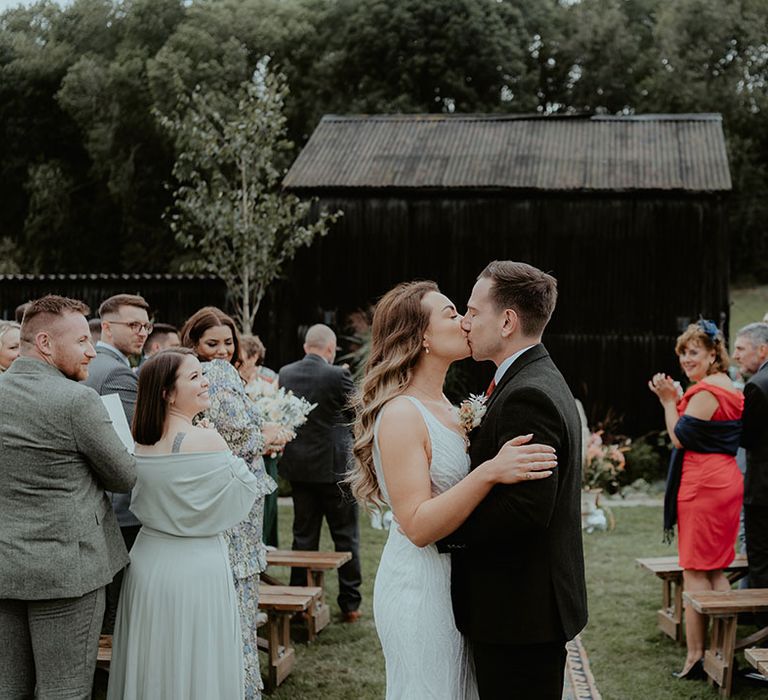 The bride and groom share a kiss at the end of the aisle as their guests applaud after the outdoor humanist wedding ceremony 