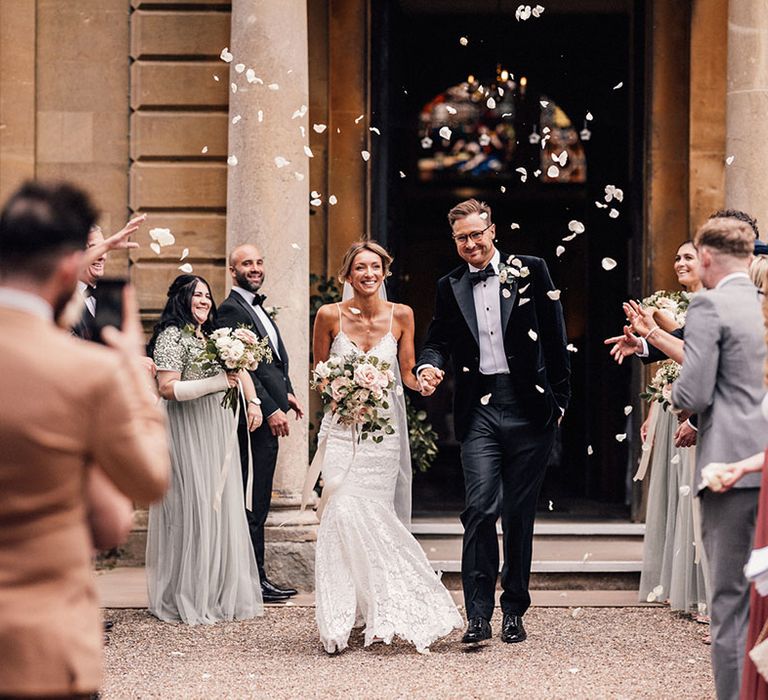 The groom in a black tuxedo walks with the bride in a fitted lace wedding dress for their classic petal confetti exit at Hanley Hall Worcestershire 