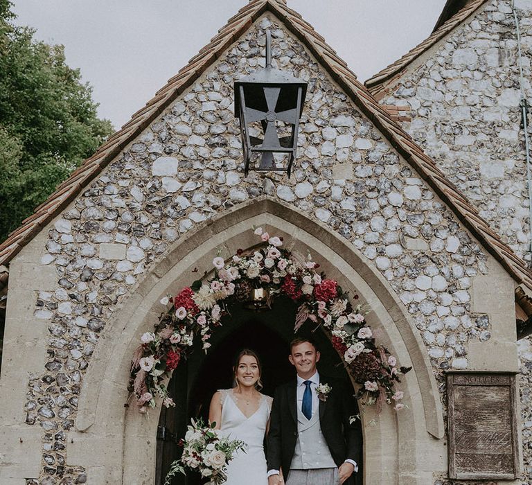 Bride & groom stand in the entranceway of church surrounded by floral arch 