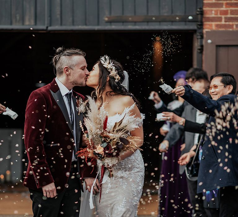 The groom in a velvet red suit jacket kissing the bride in a lace Zavana bridal wedding dress for their confetti exit 