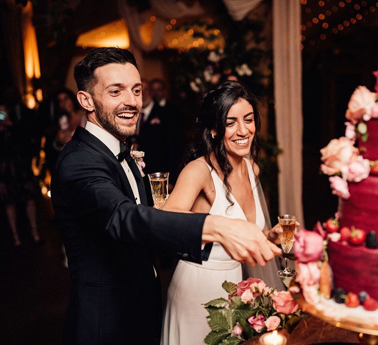 The bride and groom cut their three tier dark red wedding cake decorated with pink peonies and roses and strawberries and blueberries 