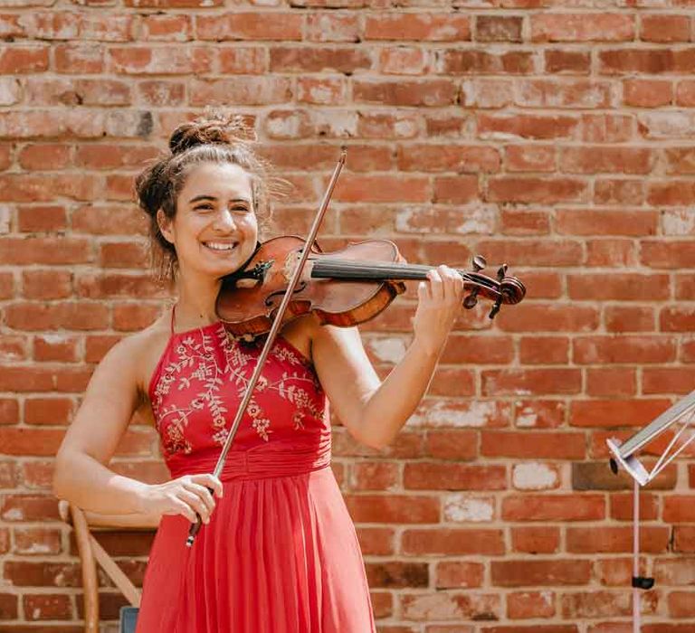 Wedding entertainer wearing red halterneck dress playing live acoustic violin at Gujarati wedding ceremony at High Billinghurst Farm