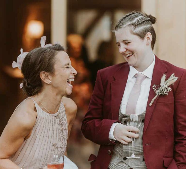 Bride in maroon bridal suit with grey waistcoat, dusky pink tie and autumnal dried flower boutonniere laughing with the mother of the bride in light pink sleeveless dress 