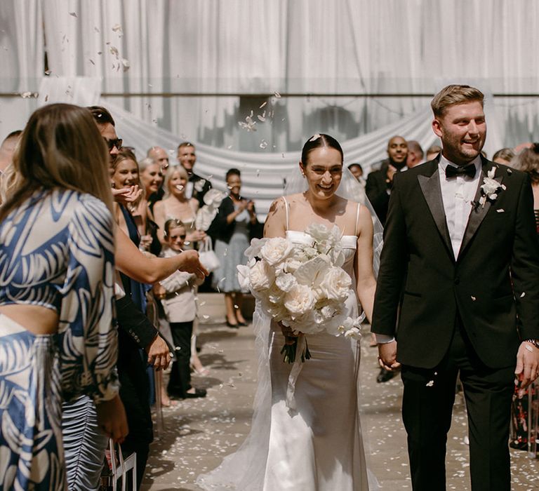 Groom in black tuxedo walks back down the aisle with the bride as white confetti is thrown over them 