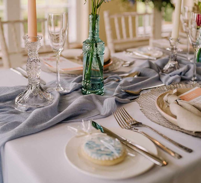 Blue glass bottle wedding centrepiece with dried spring florals, tapered candle in glass candlestick holder and cornflower blue wedding table runner 