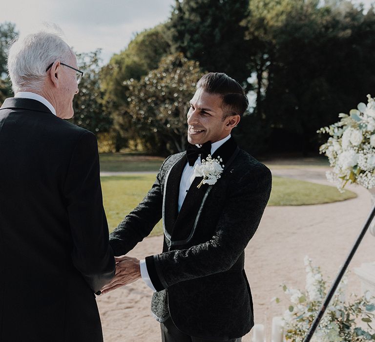 Groom in black tuxedo with black bow tie and white carnation and dried flower boutonniere with father of the bride 