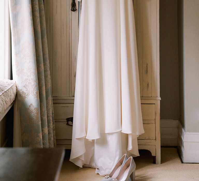 Bride's Andrea Hawkes wedding dress hanging on the wardrobe with pale blue block heel shoes 