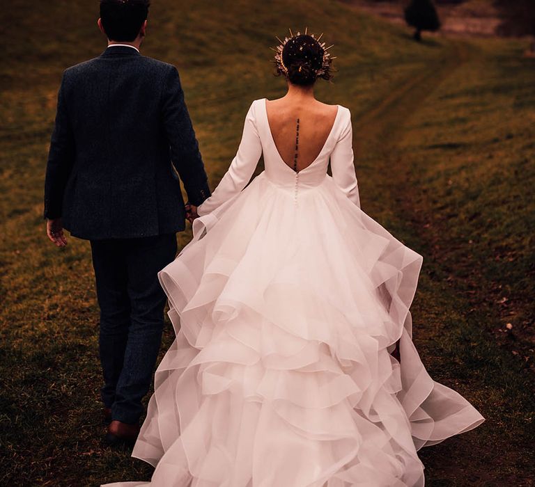 Bride in long sleeve ruffle gown with low back walking with the groom in navy suit 