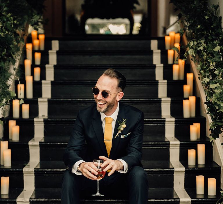 Groom in navy suit with yellow tie with yellow buttonhole and sunglasses groom accessories drinking glass of wine 