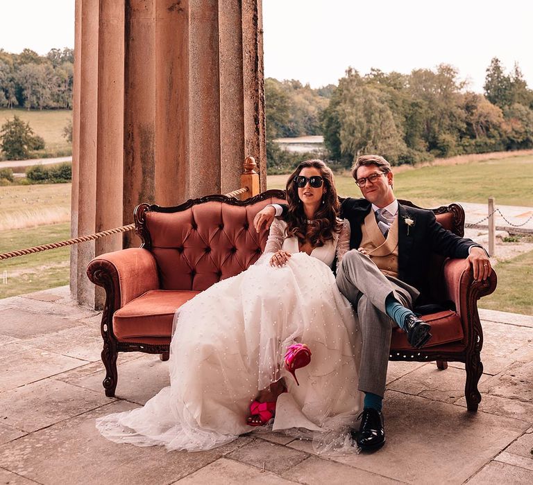 The bride and groom sit outdoors at their mansion wedding with bride wearing hot pink wedding shoes 