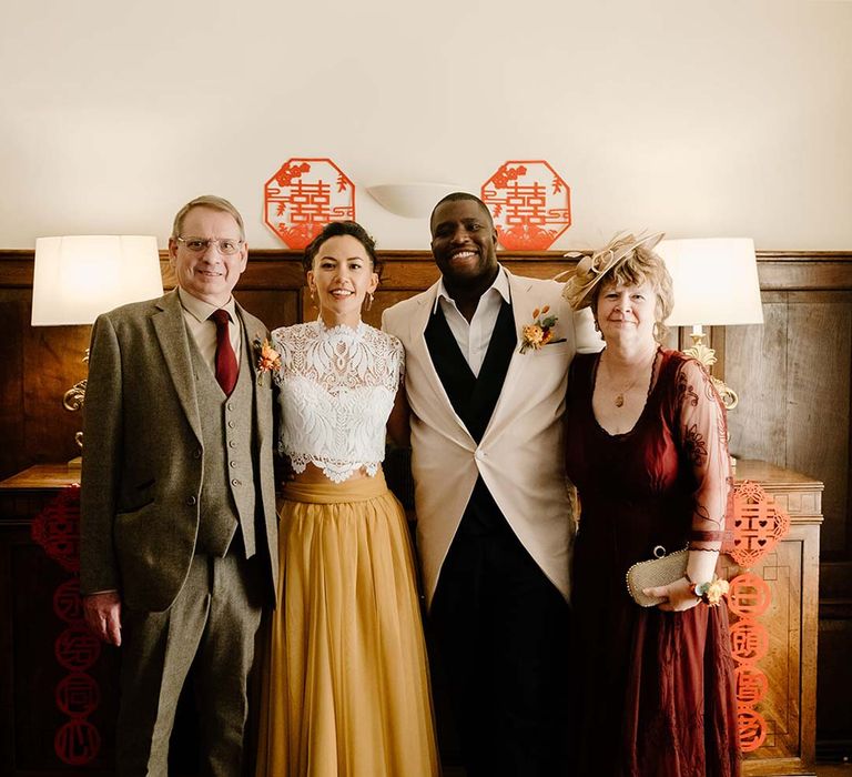 Groom incream morning suit with forest green grooms waistcoat and trousers, forest green pocket square and orange boutonniere standing with bride in white mesh lace bridal crop top and yellow wedding skirt and wedding guests during Chinese Tea Ceremony at Broadfield Court