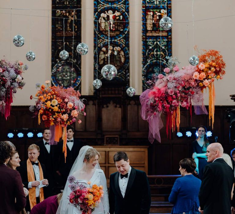 Bride in asymmetrical off shoulder long sleeve satin wedding dress with cathedral length personalised veil holding colourful bridal bouquet with pink garden roses, purple giant alliums, dark red poppies and foliage with groom in velvet tuxedo and black bowtie with puppy at Fabrica Brighton wedding venue with suspended colourful flower arrangements and suspended disco ball wedding decor 