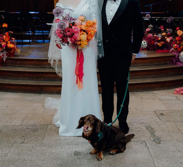 Bride in asymmetrical off shoulder long sleeve satin wedding dress with cathedral length personalised veil holding colourful bridal bouquet with pink garden roses, purple giant alliums, dark red poppies and foliage standing with groom in  in velvet tuxedo and black bowtie with puppy at Fabrica Brighton wedding