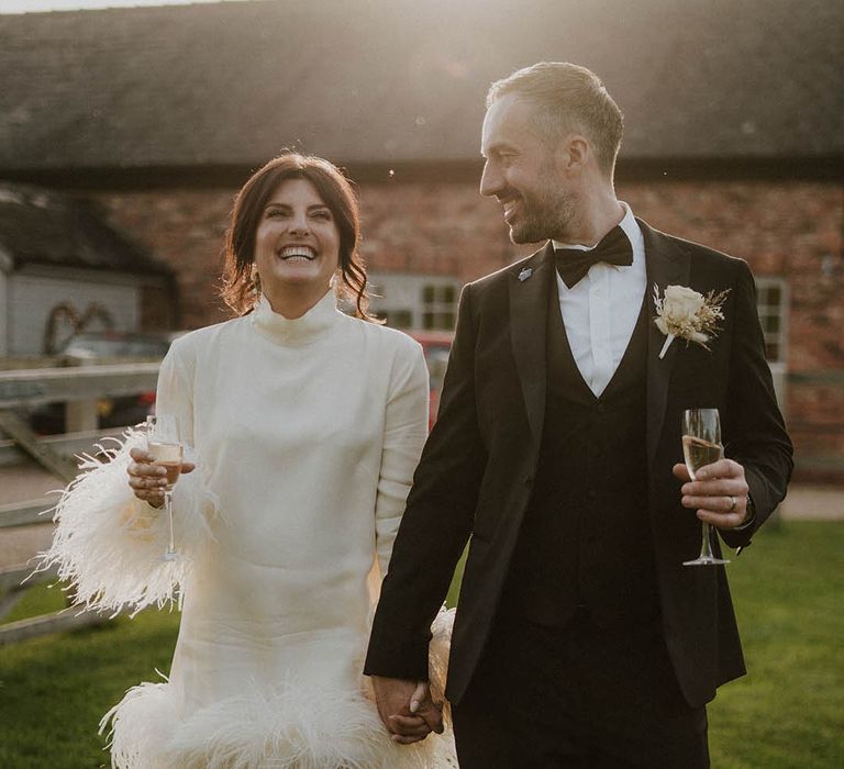 Groom in traditional black tie outfit walks with the bride in long sleeve feather wedding dress 