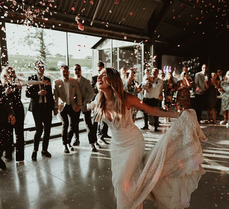 Bride wearing delicate lace wedding dress with tiara celebrating with the groom as the wedding guests throw pink and red confetti