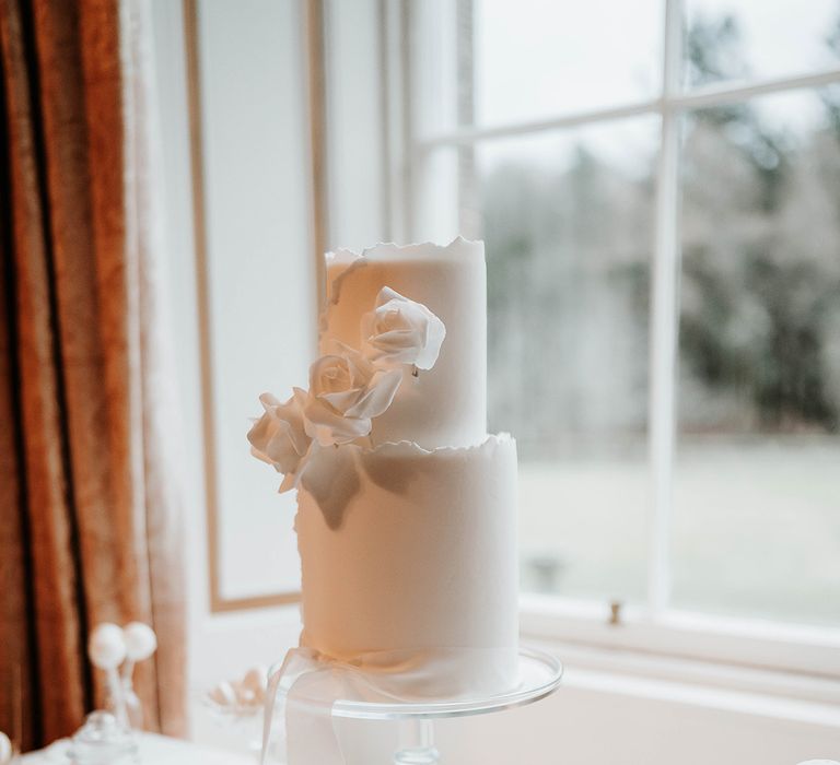 Two tier white iced wedding cake decorated with a white ribbon and white roses on glass cake stand with white cake pops, iced lollies and macarons