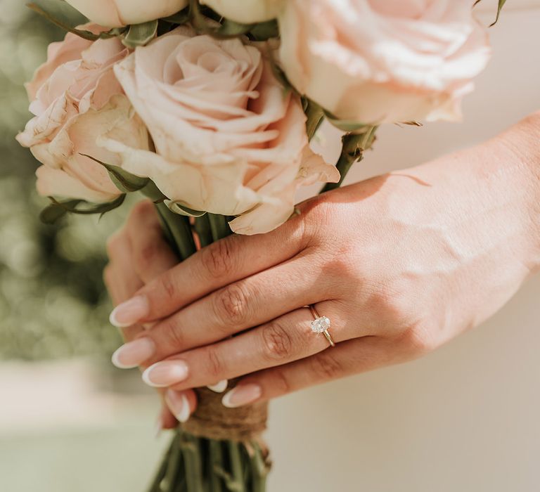 Bride wearing classic diamond oval engagement ring with French tip wedding nails holding pink rose bouquet 