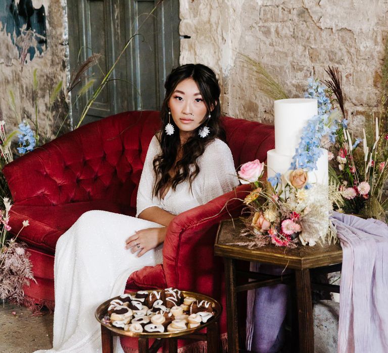 Bride sitting on a red velvet sofa next to a minimal wedding cake decorated with colourful flowers 