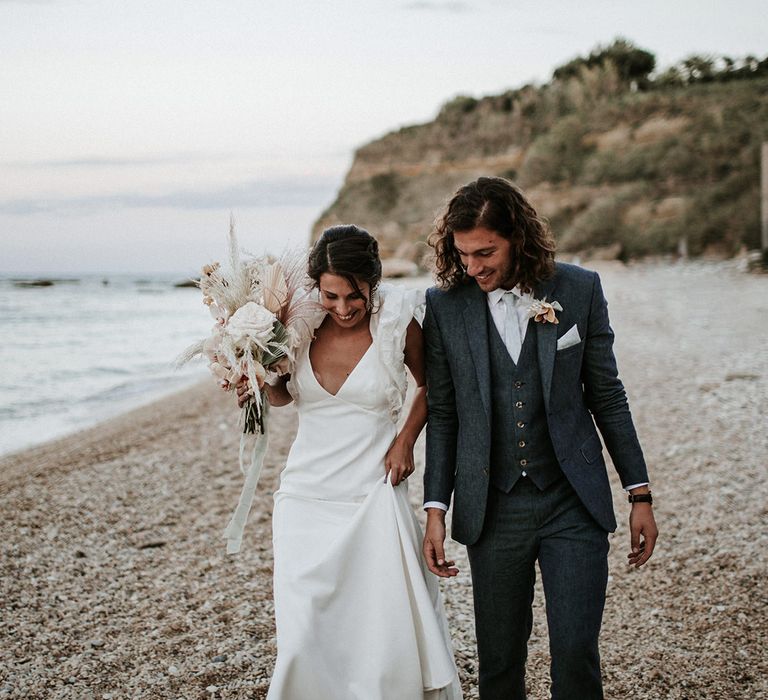 Groom in a navy blue suit walking on the beach with his bride in a Rime Arodaky ruffle sleeve wedding dress holding an orchid wedding bouquet 