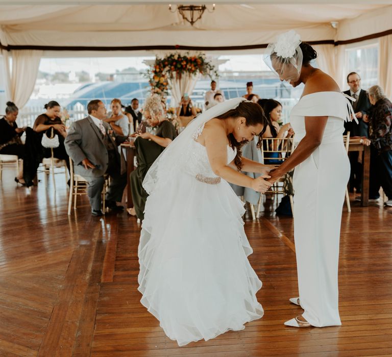 Black bride in flat wedding shoes, jumpsuit and blush veil and bi-racial bride in a strapless wedding dress with full skirt on the dance floor 