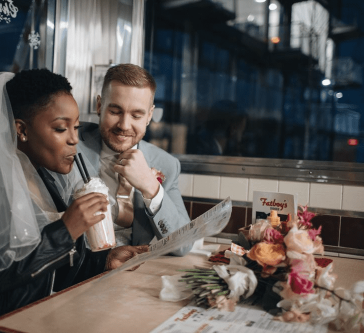 A bride and groom sit at a table in a cafe. The bride drinks a milkshake. She has cropped hair and wears a veil with leather jacket. Wedding hairstyles for Black brides.