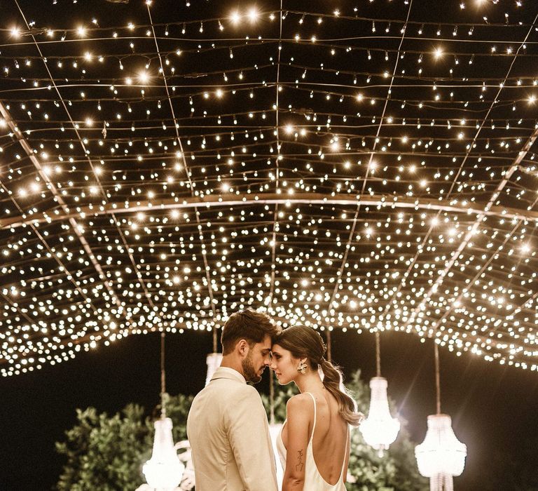 Boho bride and groom standing under a canopy of fairy lights in a beige suit and slip wedding dress