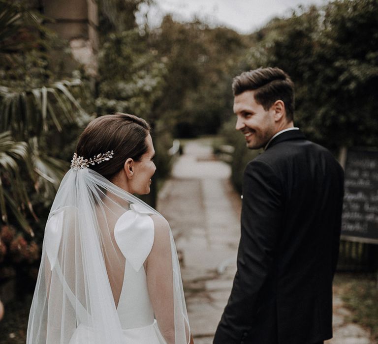 Bride in white Jesus Piero gown and cathedral length veil holds hands with smiling groom in black Tiger of Sweden tux in gardens at German wedding