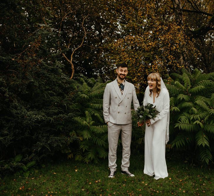 Portrait of the groom in a grey suit with patterned shirt and the bride in a jumpsuit with appliqué veil 