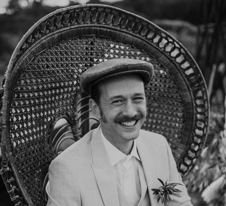 Black and white portrait of the Franco-American groom in a Peaky Blinders wedding suit and cap sitting on a Peacock Chair 