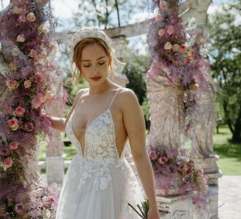 Bride wears low cut gown with appliqué design surrounded by lilac florals