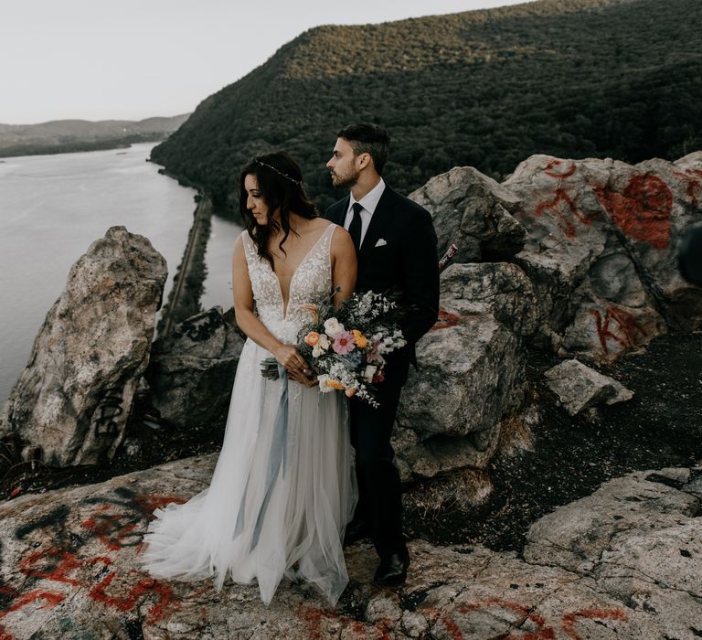 Groom embraces bride as they look across the Hudson River whilst holding colourful bouquet