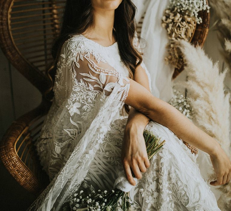 Bride in a lace Julita London wedding dress and flower crown sitting on a wicker chair with dried flower arrangements 