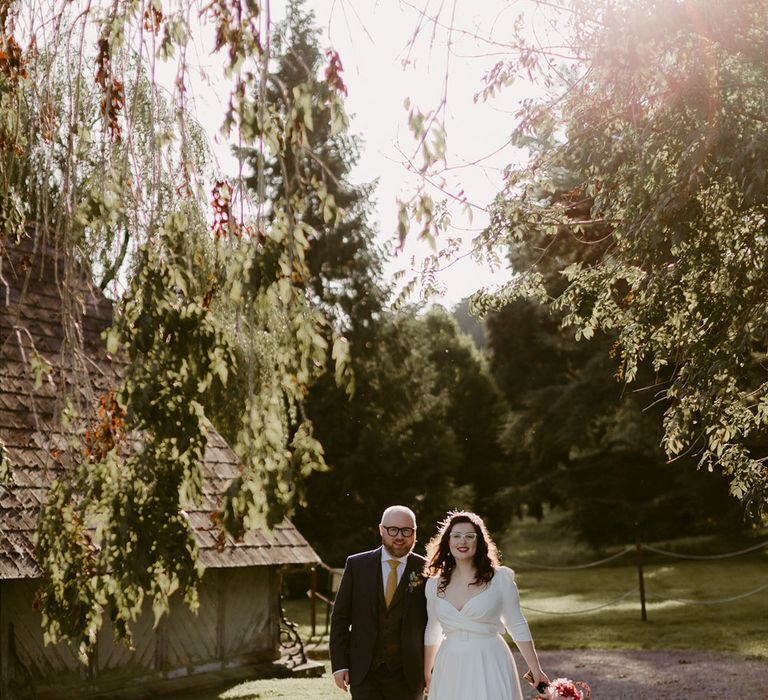 Groom in navy suit and yellow tie stands with bride in white cat eye glasses and bridal headband holding white and pink rose and pampas grass bouquet